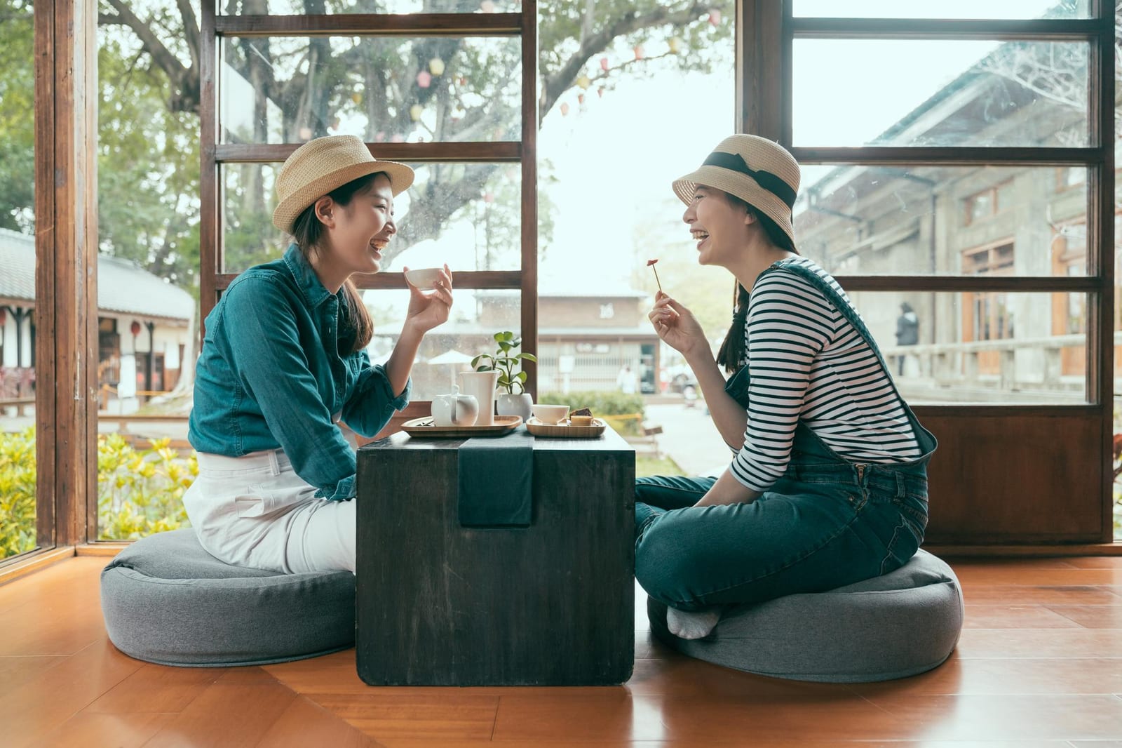 麦わら帽子をかぶった二人の女性が木の窓辺の床座席でお茶を楽しんでいる様子。